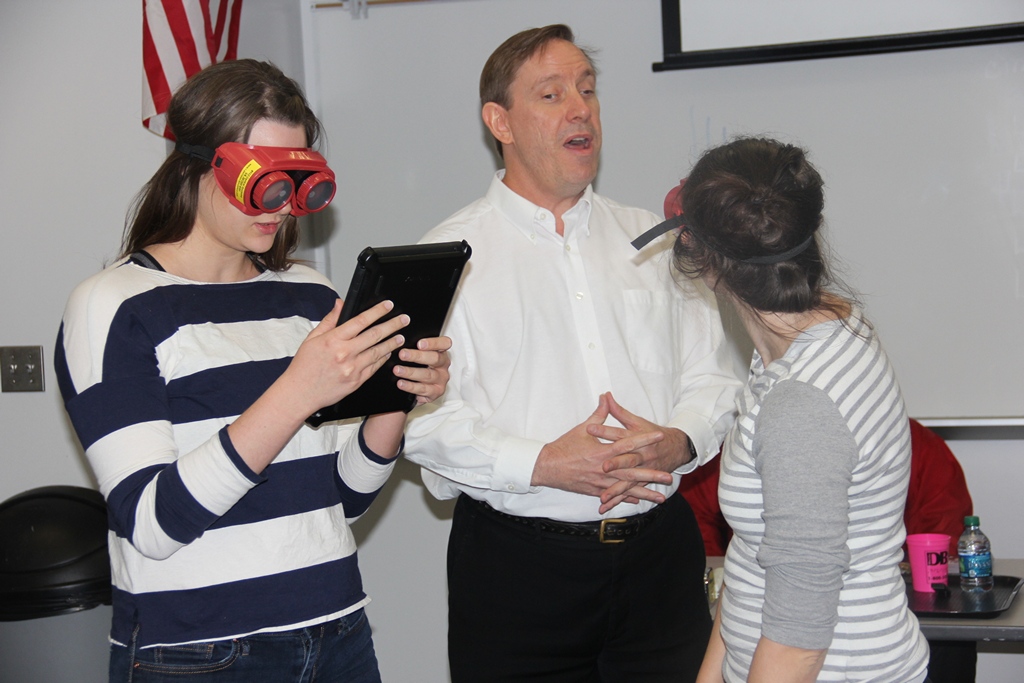 While wearing vision simulators, two young women use an iPad and are being assisted by a man in a white shirt.